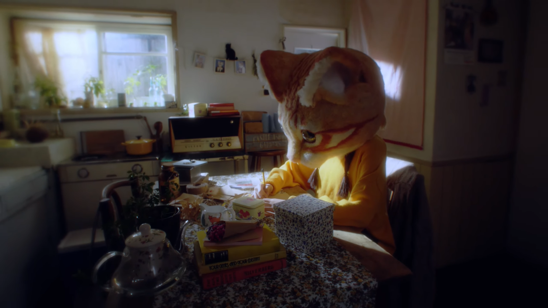 Hyunjin sitting at a table and writing on a piece of paper. She is wearing a large orange cat head.